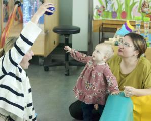 Toddler in pink dress reaches up to a ball held by a woman in a striped sweater while another woman smiles