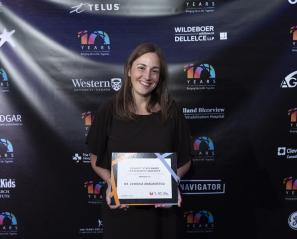 Dr. Evdokia Anagnostou stands holding her framed award, smiling. She is in front of a backdrop with various logos from Western, SickKids and other organizations.