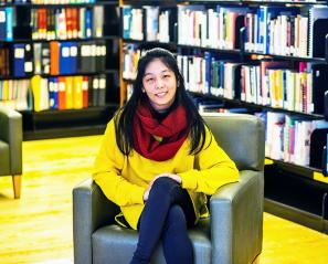 Youth leader Lexin sits in a chair surrounded by shelves of books. She is wearing a yellow sweater and a red scarf.