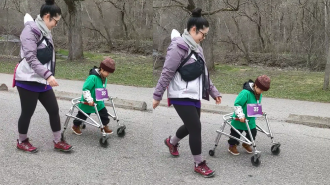A young child walking on pavement. He is using a walker and is wearing a green t-shirt, brown shoes and a red beanie hat. His mom is walking beside him. She is wearing her hair in a bun, a purple and white rain jacket and black athletic pants.