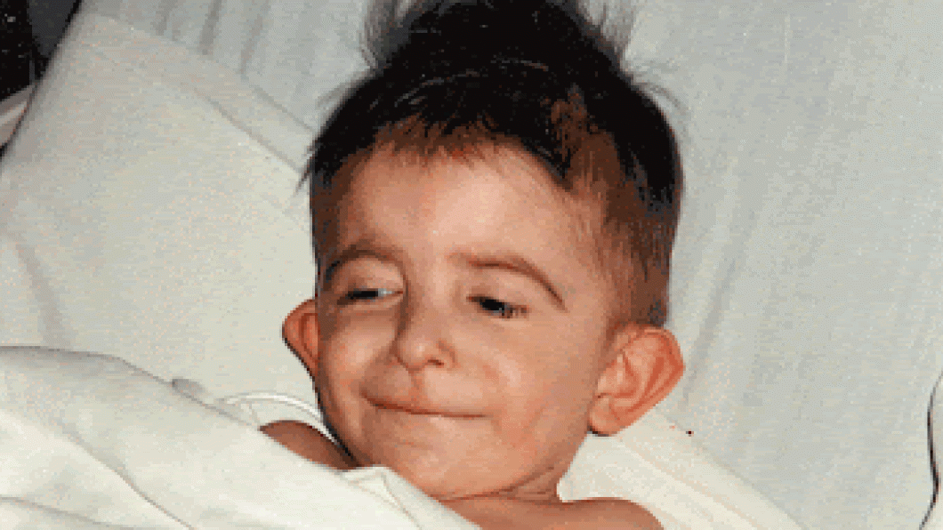 Small boy with brown hair smiles under sheet of a hospital bed
