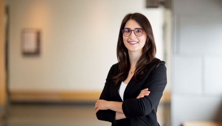 A woman with brown hair and glasses wearing a black blazer with a white t-shirt with their arms crossed.