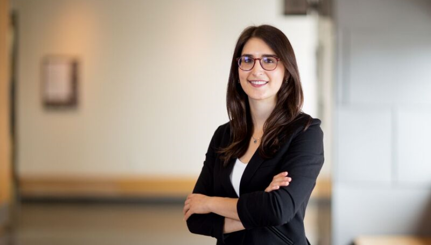 A woman with brown hair and glasses wearing a black blazer with a white t-shirt with their arms crossed. 