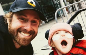 Man in baseball cap smiles beside young girl in stroller wearing nasal prongs