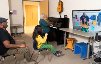 Child in yellow shirt sits in front of screen with icons with a woman on piano beside her and a man sitting behind her