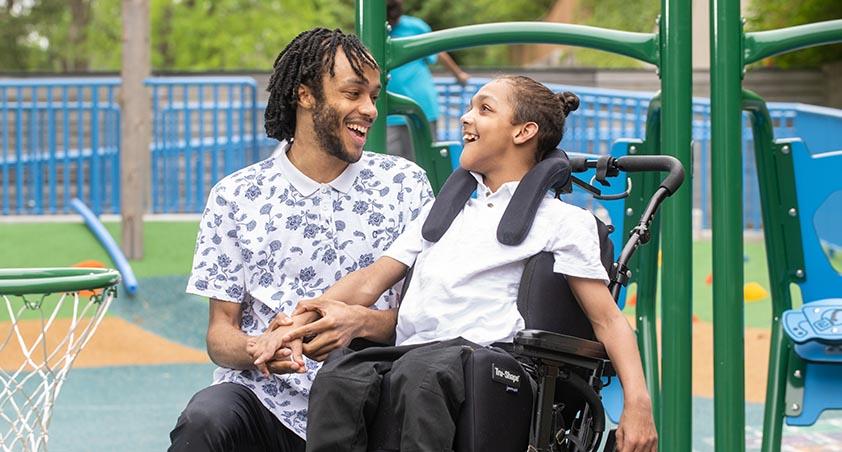 Two teenagers playing in a playground setting. One teenager sitting on a wheelchair