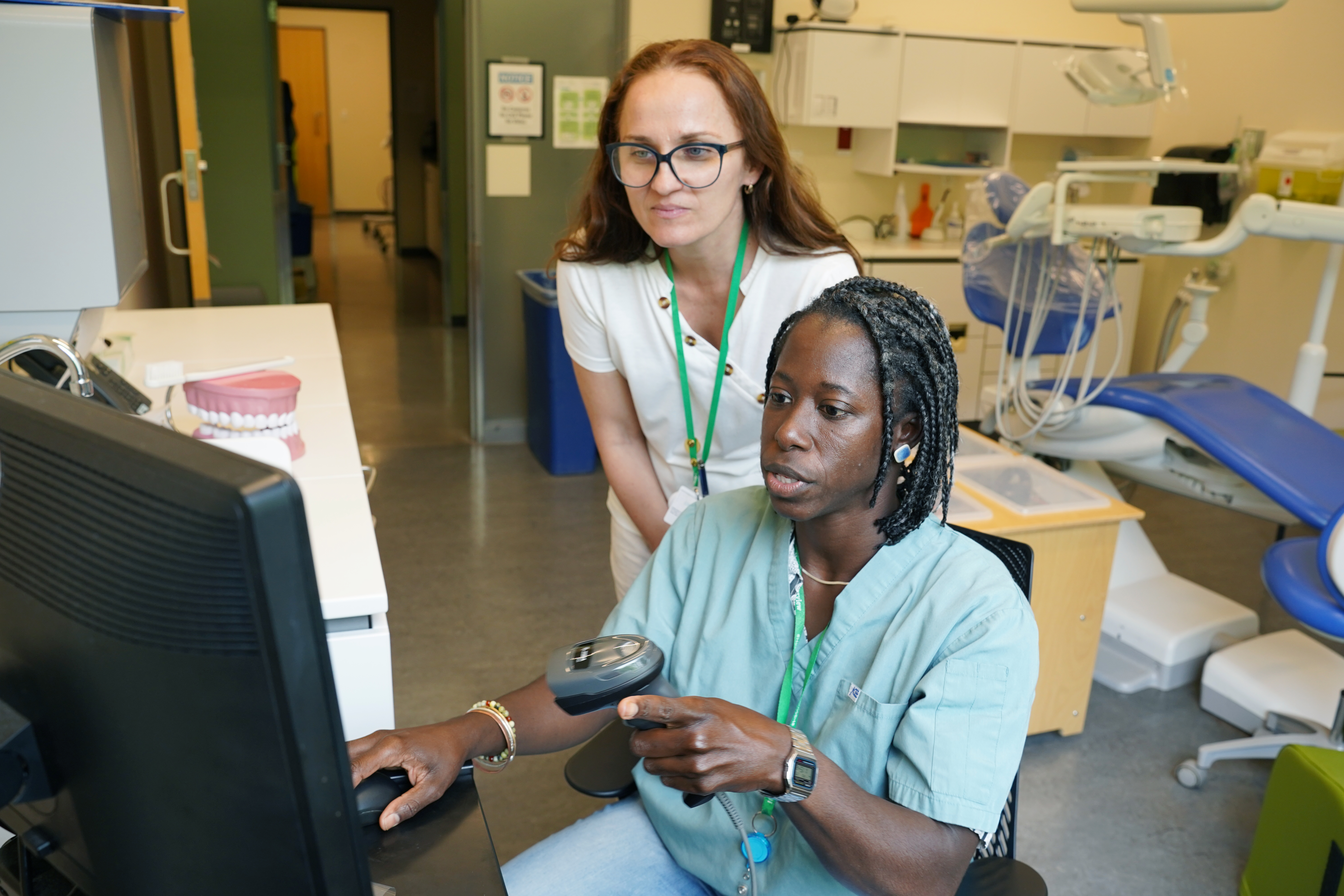 two dental professionals in a clinic setting, with one seated using a computer and holding a dental model while the other stands observing.
