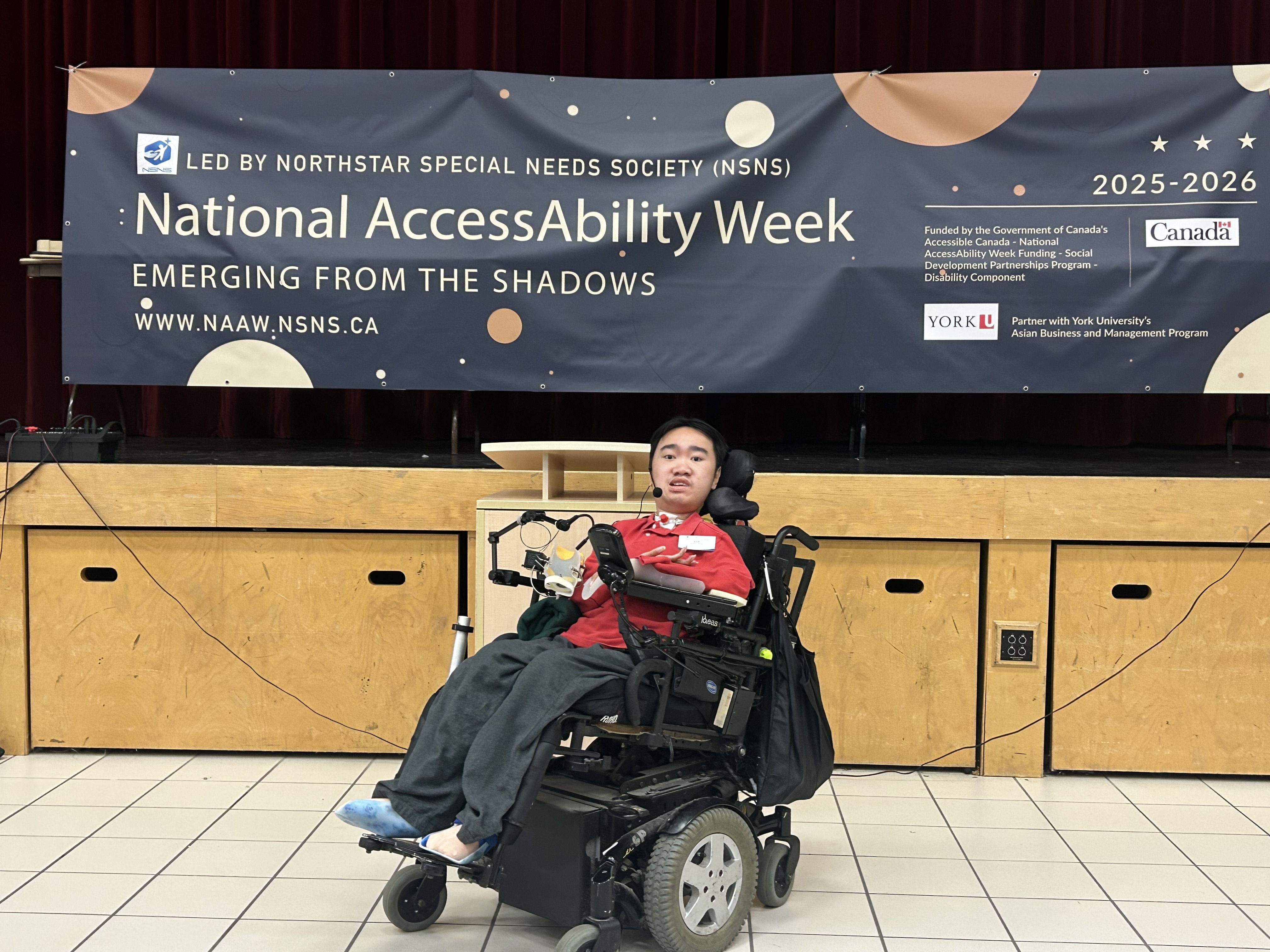 Jeffrey, who uses a powerchair, poses for a photo in front of a stage, below a banner that reads: 'National AccessAbility Week'