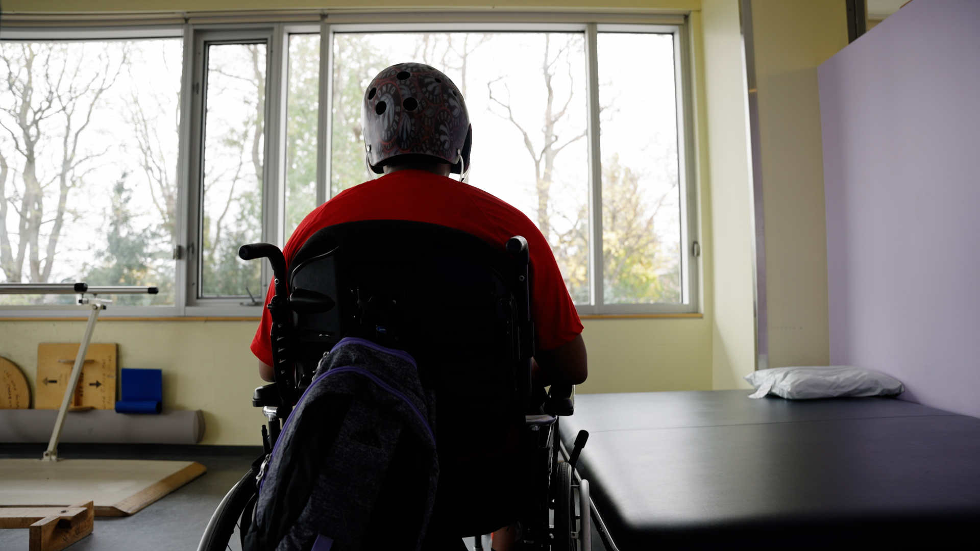 Photograph showing a person wearing a helmet sitting in a wheelchair facing large windows in a rehabilitation or therapy room. The room includes a padded therapy table, exercise equipment, and natural light highlighting a calm, supportive environment.