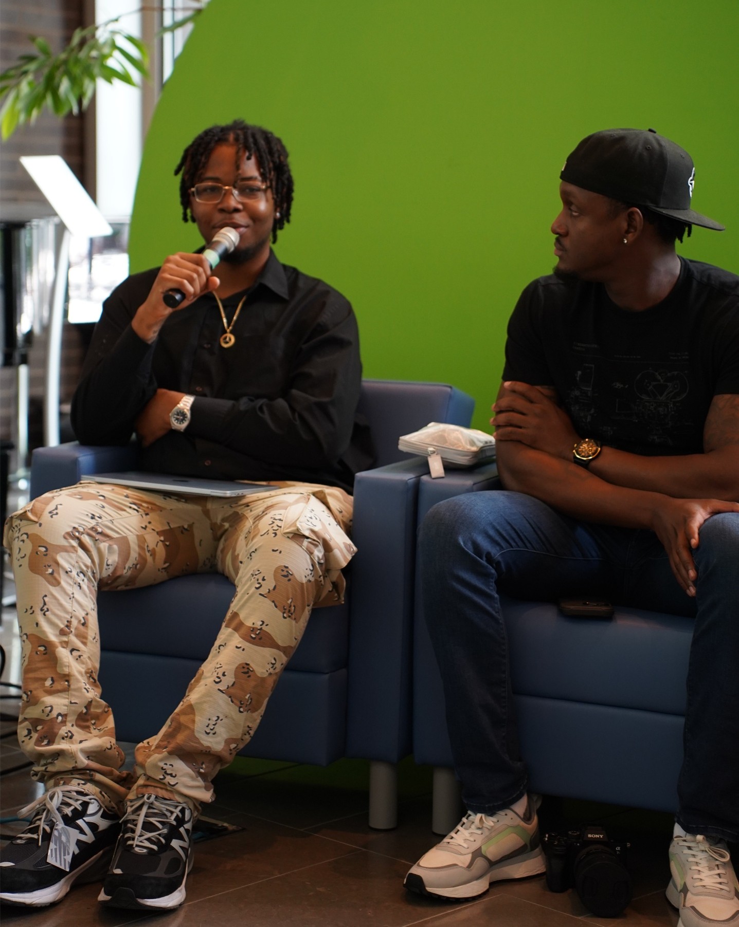 Photograph of two men sitting on blue chairs against a green wall, engaged in conversation. One man holds a microphone and wears patterned pants with sneakers, while the other wears a black shirt, dark pants, and a backward cap.
