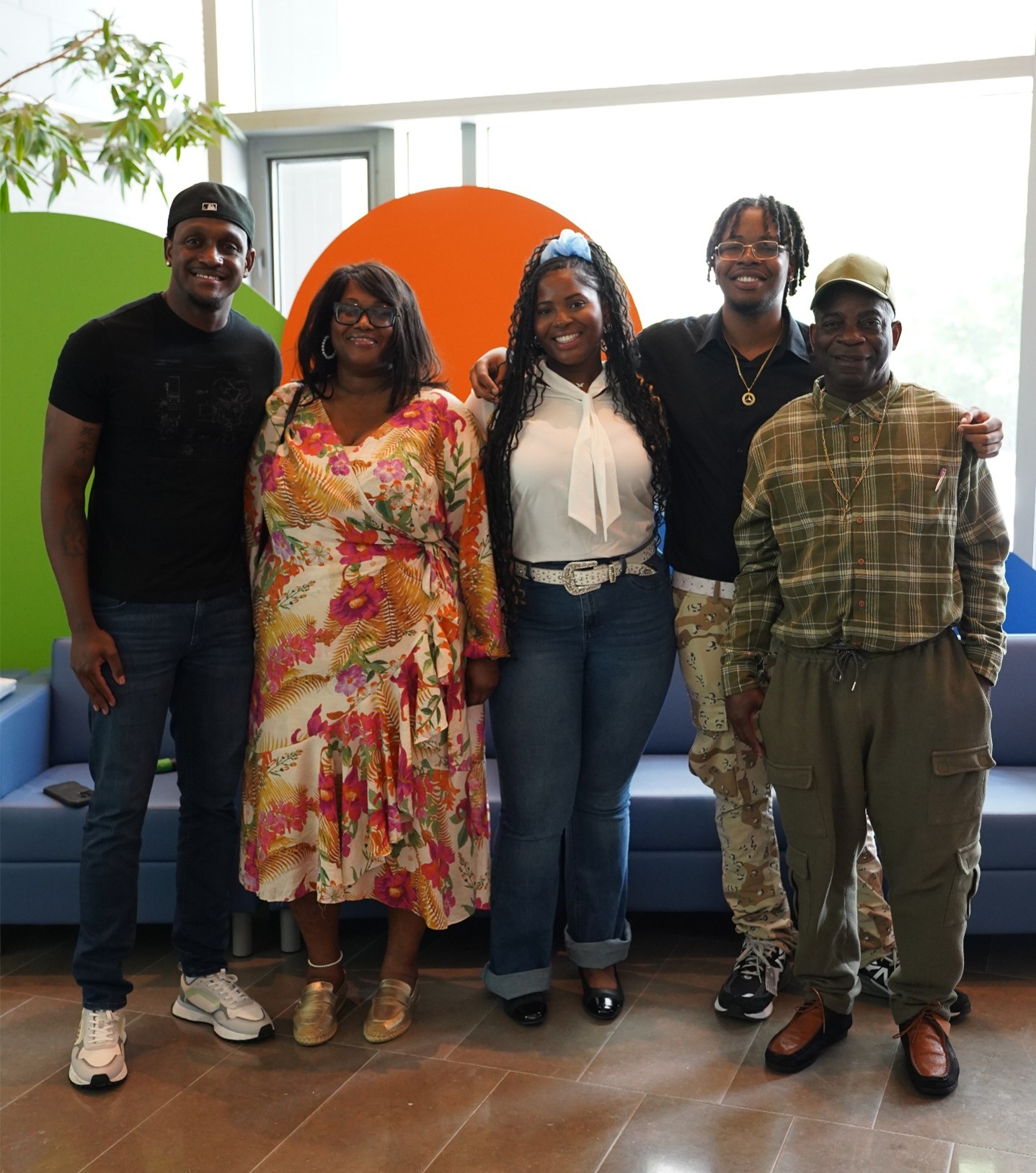 Photograph of five people standing indoors in front of colorful circular wall decorations in green, orange, and blue. Group includes individuals wearing casual and business attire, with a plant visible on the left and seating furniture in the background.