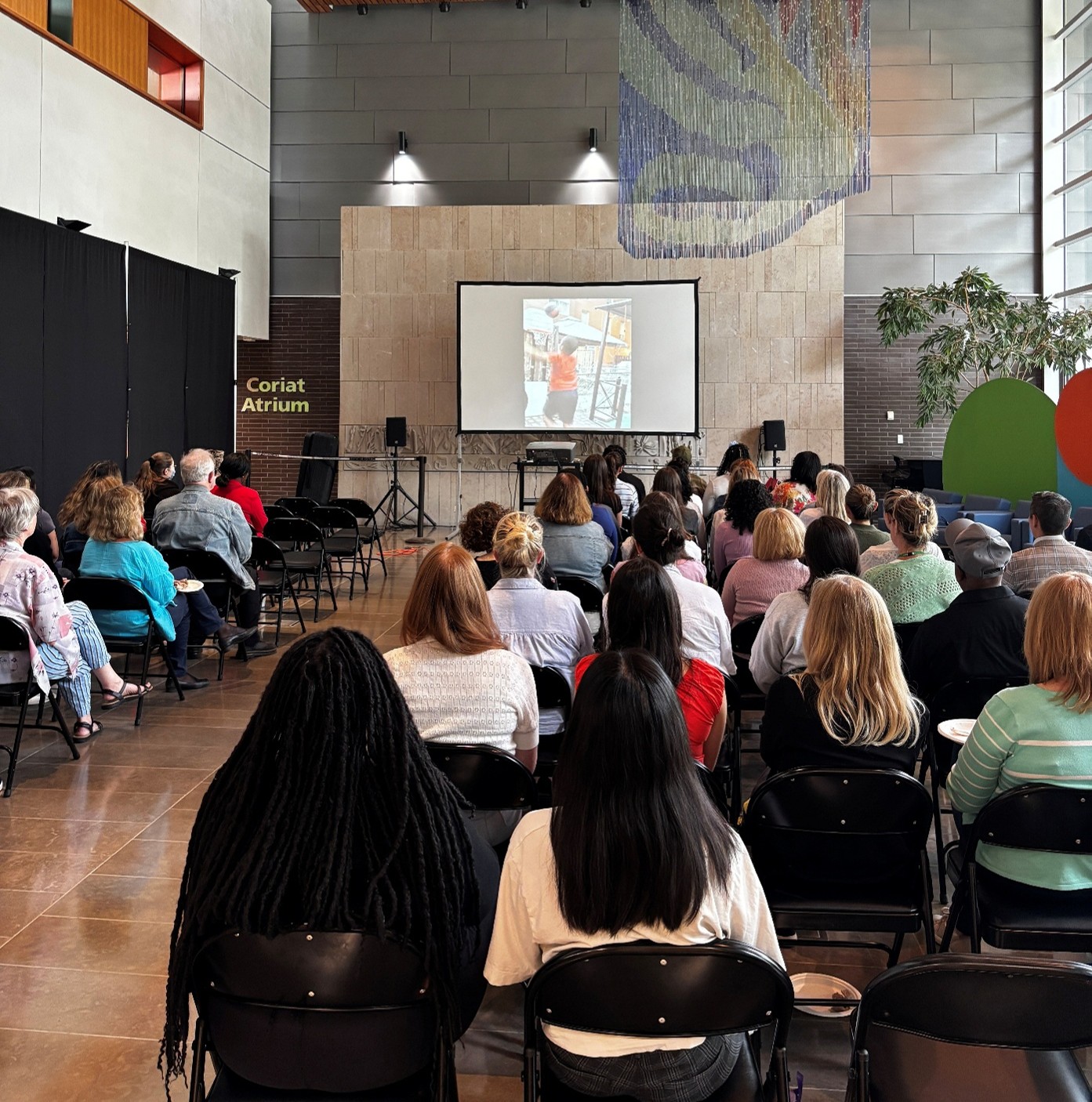 Photograph of an indoor presentation with an audience seated facing a screen displaying a slide. The setting features a high wooden ceiling, stone walls, and modern art installations, including a large hanging sculpture and a spider figure on the left.
