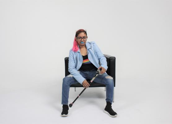 Brown woman wearing a rainbow shirt and jeans with pink ponytail and cane