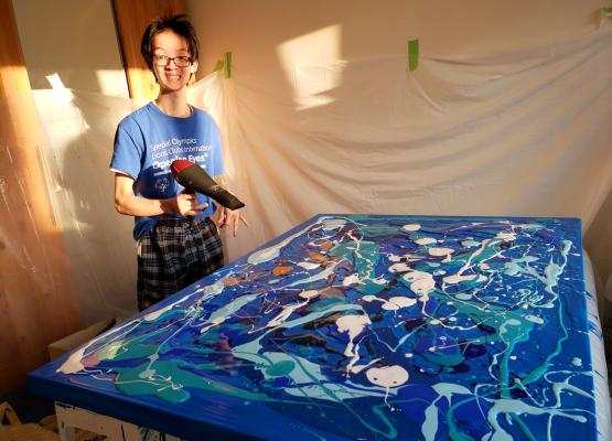 Teenage boy beside canvas with blue shades of paint poured over it holding blow dryer