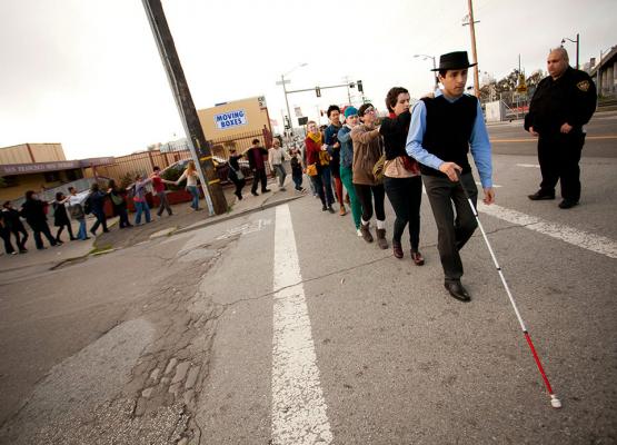 Man with a white cane leading a line of people along a city street