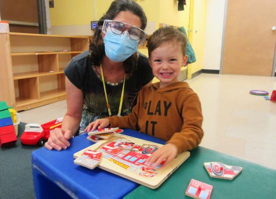 Woman with mask and face shield and goggles beside preschool boy doing a puzzle