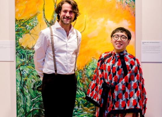 Bruce and Sean smile together at the opening reception for Through A Tired Eye. Sean is wearing a red kimono-like dresshirt with red fringes. Bruce wears a white dress shirt, black pants with grey suspenders. Behind them is an acrylic painting of greenery with a yellow skyline.