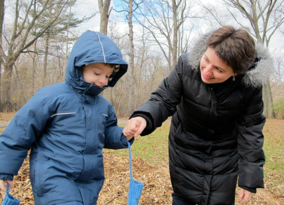 Woman holds hand of boy wearing blue winter coat with mittens on strings