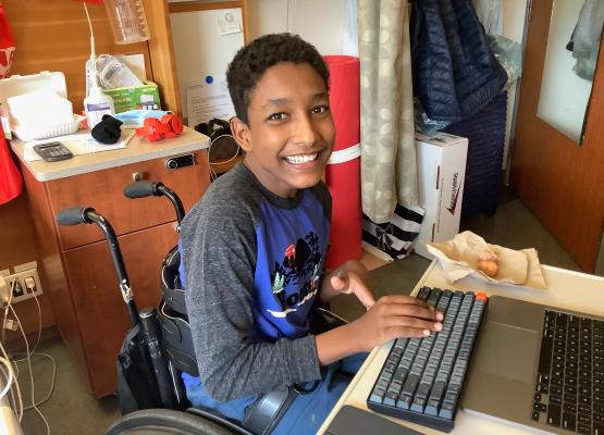 Teen with wide smile sits in wheelchair at computer in hospital room