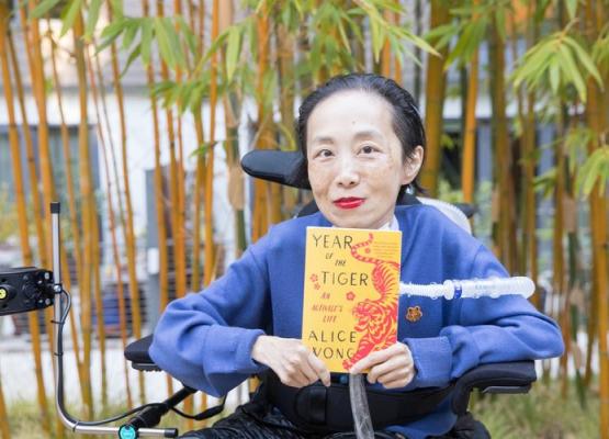 Woman with red lipstick and blue shirt holds orange book in wheelchair