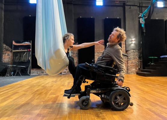 A woman in a silk hammock reaches her arm out to a man in a power wheelchair
