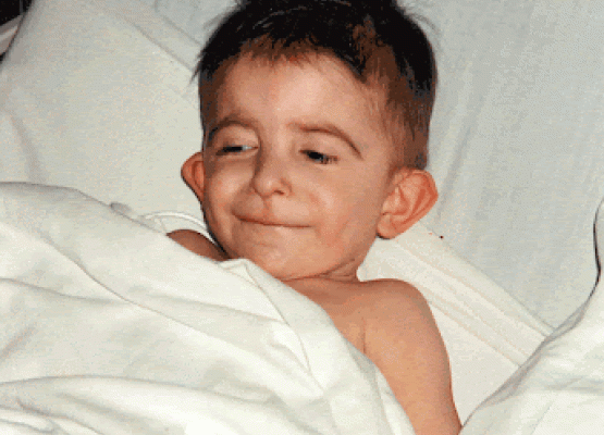 Small boy with brown hair smiles under sheet of a hospital bed