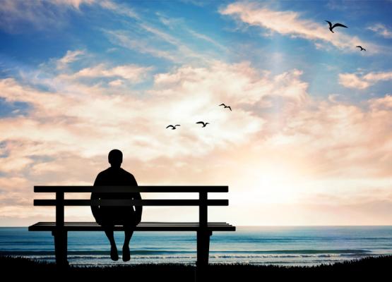 Silhouette of man alone on bench looking out to sea and clouds and seagulls.