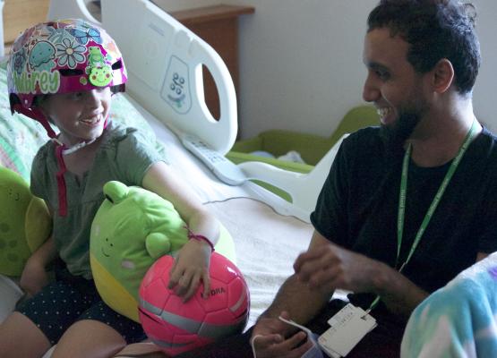Girl with brightly coloured helmet and holding stuffies smiles on hospital bed at man in black shirt and beard who is also smiling