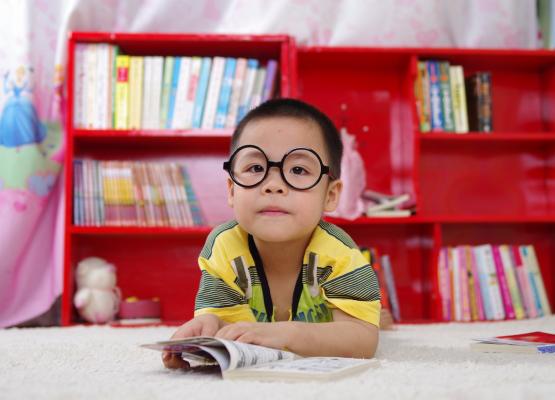 Boy in yellow shirt with big black reading glasses rests on arms on floor with red bookcase behind.