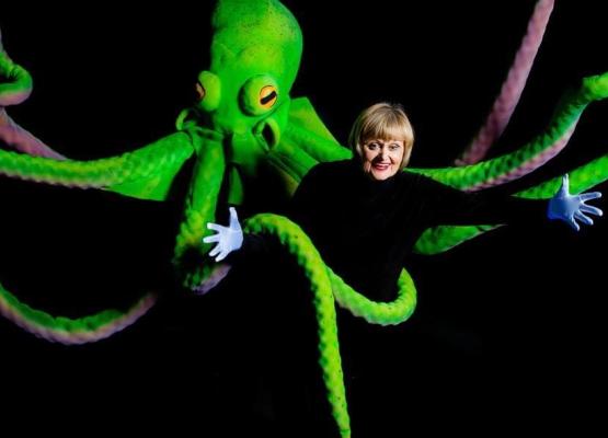 Woman in black smiling surrounded by giant bright green octopus and its tentacles.