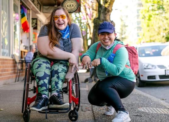 Two young women, one in a red wheelchair with lucky clover leggings, the other crouched down beside her in a blue baseball cap