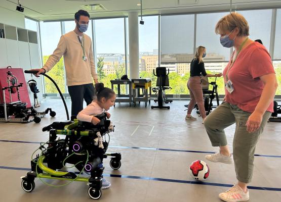 Small girl in green robot-powered walker faces therapist who is kicking a soccer ball