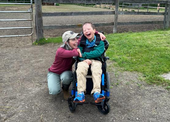 Woman with blonde hair and baseball cap crouches down beside boy in green sweatshirt in wheelchair both smiling 