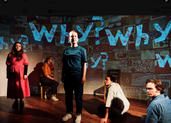 Actors standing and sitting on a stage with the word 'Why?' superimposed in blue on a screen behind them