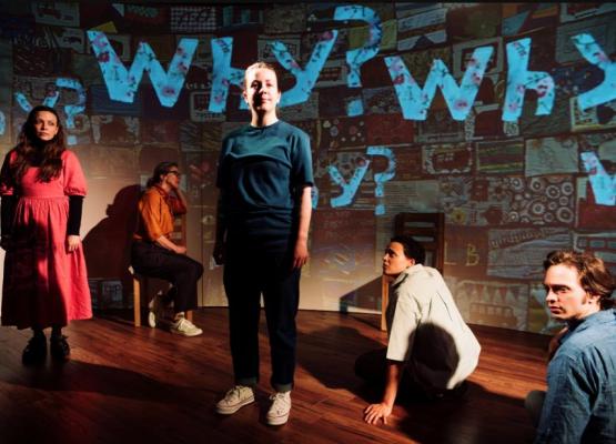 Actors standing and sitting on a stage with the word 'Why?' superimposed in blue on a screen behind them