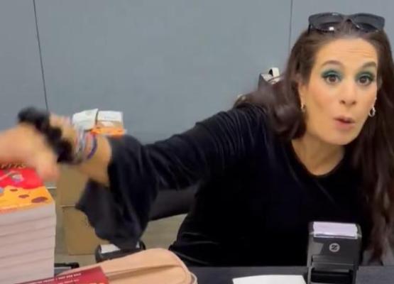 Woman with long dark hair sits at desk and puts hand on a pile of brightly coloured books