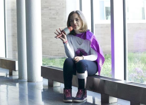 Girl with shoulder length brown hair and pink microphone wears purple cape and sits on bench