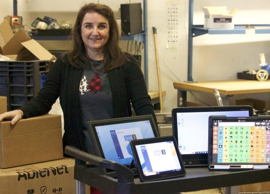 Woman with long hair stands in front of trolley with a number of computers displayed and boxes behind in warehouse