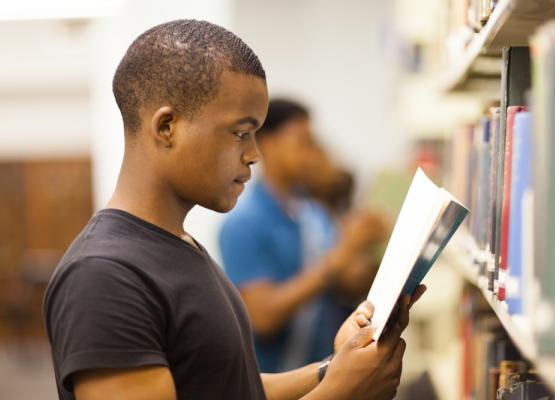 Young man in black t shirt with close cropped hair reads a publication in a library