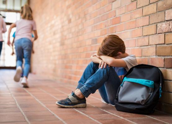 Young boy with head in hands in school hall as other kids run away