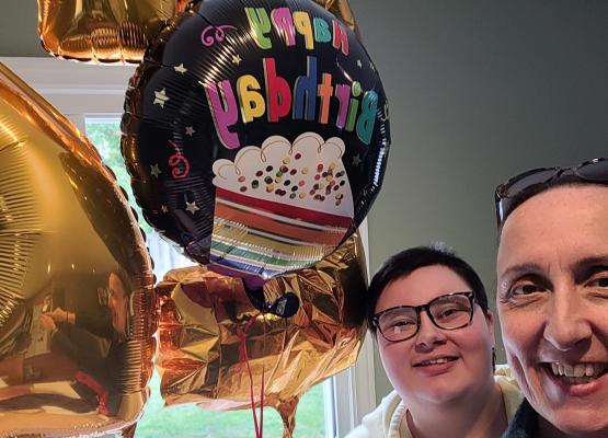 Two women's faces smile beside brightly coloured birthday balloons