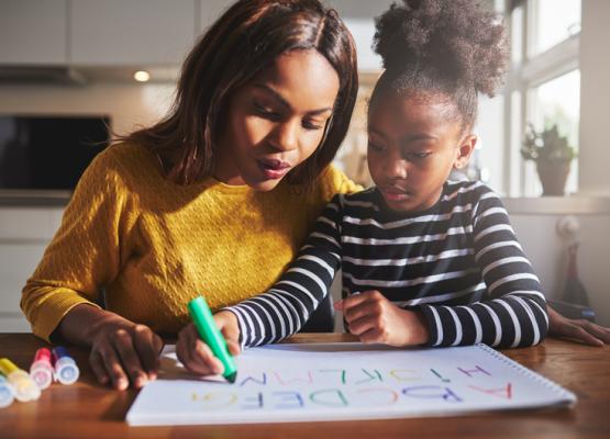 Woman in yellow sweater helps daughter in striped sweater write out alphabet