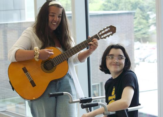 Woman stands playing guitar beside girl sitting on walker smiling