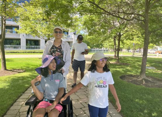 Girl holds hand of teen in wheelchair on path in the park with two people behind them