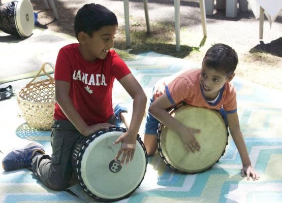 Boy with dark hair and red Canada shirt and boy with dark hair and orange shirt play drums