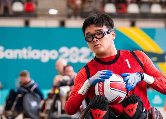 Man with dark hair and glasses wearing red athletic top and gloves holds ball in wheelchair