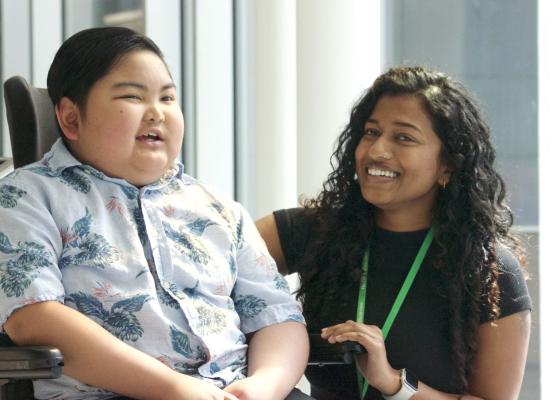 Boy with dark hair smiles in wheelchair beside woman with long dark hair who is also smiling