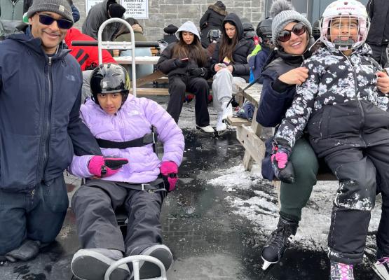 Teen in purple jacket on sledge holds hand of man beside girl in skates sitting on woman's lap