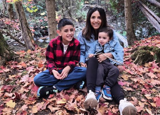Two boys with dark hair sit in lap of woman with dark hair among colourful fall leaves