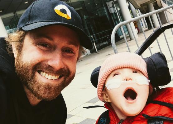 Man in baseball cap smiles beside young girl in stroller wearing nasal prongs